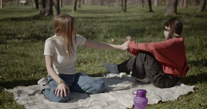 Girls enjoy a day in the park, sitting on a blanket and playing around. They laugh, tickle each other, and have a genuinely fun time together, enjoying each other's company and the pleasant weather.