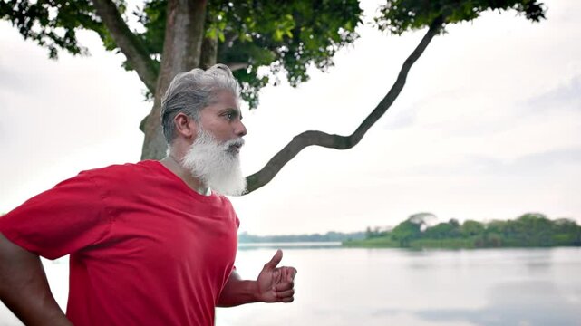 Mature Indian man with a beard jogging in a lakeside park In Singapore. Slow motion.