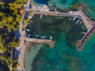 High-resolution drone image showing a small man-made harbor with several boats moored around the perimeter. Crystal-clear turquoise water reveals underwater details and rocks. 