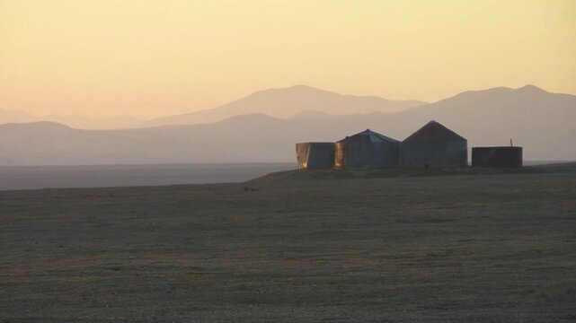 Old silos barely lit by the last rays of the setting sun stand amidst a vast barren field in California's Carrizo Plain.  Layered mountain ridges recede into the distance under a sepia sky. 