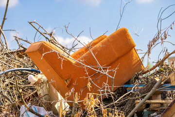 A worn out orange chair abandoned in a dump in a pile of branches, trash and debris against a...