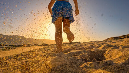 LOW ANGLE VIEW, LENS FLARE: Sporty woman in skort on an evening run along a sandy ocean shore. At sunset, she runs barefoot on beach in blue sportswear, leaving flying sand behind in golden light.