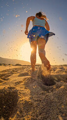 LOW ANGLE VIEW, LENS FLARE: Sporty young woman in blue skort runs barefoot across a sandy ocean beach during golden sunrise, kicking up sand as the warm morning sunlight backlights her silhouette.