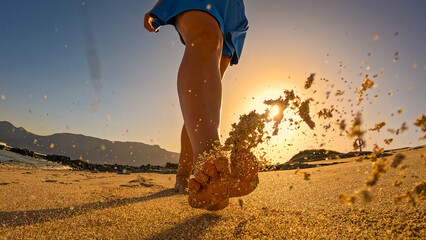 LOW ANGLE VIEW, LENS FLARE: Young lady in blue skort during a walk along sandy shore in golden light. Vacationer is walking barefoot and enjoys carefree summer moments on holidays at Canary Islands.