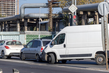 A small accident with three cars colliding with each other, on a city road, with an industrial building and pipes visible in the background.