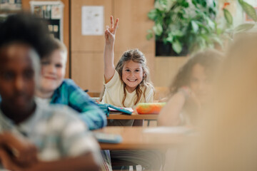 Smiling schoolgirl raising hand and showing peace sign in classroom