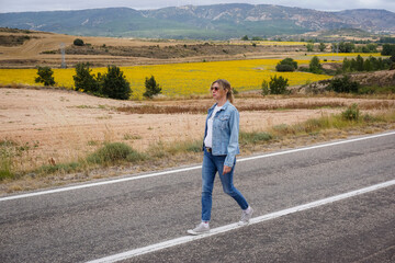 Young blonde woman wearing a jacket and blue jeans walking along a deserted road