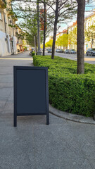 A blank black signboard stands on a sidewalk surrounded by neatly trimmed bushes and trees. The scene is set in a sunny urban environment with parked cars in the background. mock up