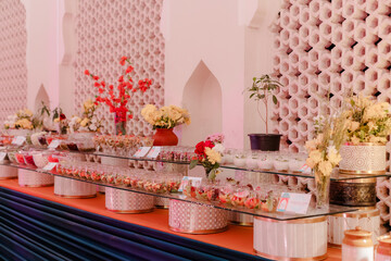 Elegant dessert buffet display with tiered glass shelves. Fresh flowers and potted plants add a touch of nature against patterned walls.
