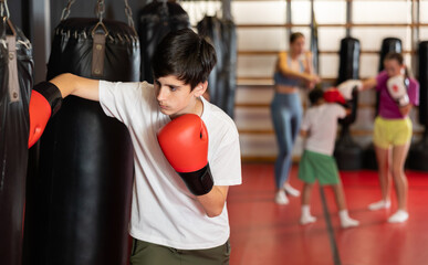 Teen boy in boxing gloves hits punching bag in gym. Playing sports develops mind and body..