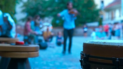 Street musician performing violin near large drum in blurred town square, capturing vibrant outdoor musical moment with artistic defocused background
