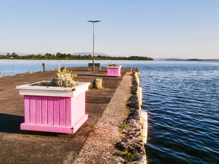 Wooden flower bed painted in pink and white is on a pier with stunning nature scenery and dramatic sky in the background. Beautiful nature. Blue water surface. Nobody. Calm and relaxed mood