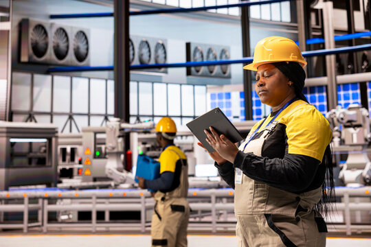 Female engineer in smart manufacturing plant monitors production line with tablet. The scene highlights industrial automation with robotic arms and solar panels, ensuring optimal factory performance.