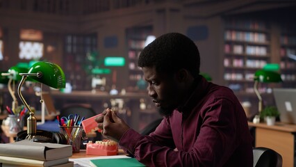 African american student examining fake teeth for dentistry thesis, working on stomatology research with appropriate tools in the campus library. Guy preparing for his career. Camera A.