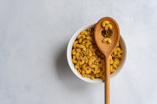 Overhead view of a bowl of raw macaroni pasta and a wooden spoon with a heart shape on a kitchen worktop