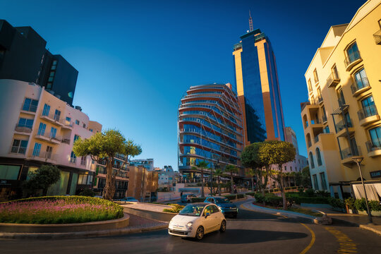 Scenic view of a white Fiat 500 car driving around a roundabout in front of the Portomaso Business Tower in Saint Julian's (San Ġiljan), Malta
