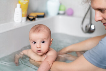 Cute baby bathing and swimming in the bath. Baby learning to swim and playing sports. Father enjoys bath time with his curious baby in a cozy bathroom. Fatherhood concept. Selective focus.