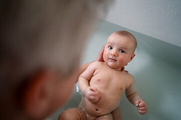 Top view cute baby bathing and swimming in the bath. Baby learning to swim and playing sports. Father enjoys bath time with his curious baby in a cozy bathroom. Fatherhood concept. Selective focus.