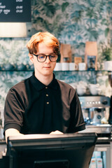 Young waiter serving customer at cash point in cafe. Man working with POS terminal. Cashier, barista checking for payment receipt. Hospitality, server and preparing a slip at the till in coffee shop