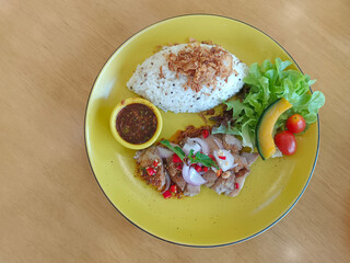 top view, Waterfall pork, fried garlic on rice and nam jim jaew in a yellow bowl on a yellow plate on a wooden background.