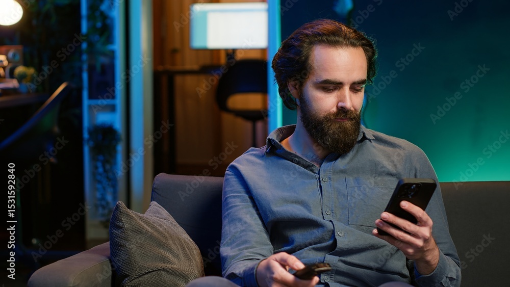 Poster smiling person on couch picking movie on streaming service using remote and texting on mobile phone.