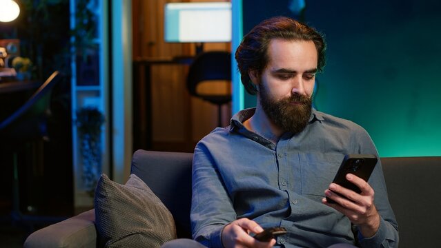 Smiling person on couch picking movie on streaming service using remote and texting on mobile phone. Happy man watching shows on television, zapping though channels, chatting on smartphone, camera A