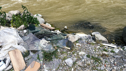 Broken glass, plastic and construction debris scattered along a rocky riverbank, polluting the natural environment and threatening the ecosystem, pollution concept