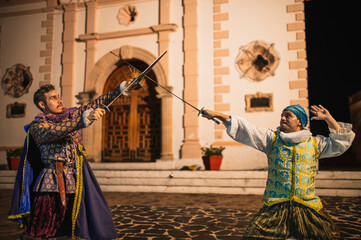 Spanish men in traditional colonial clothing dueling with swords called foils and daggers in a...