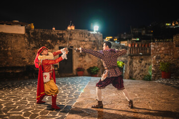 Spanish men in traditional colonial clothing dueling with swords called foils and daggers in a...