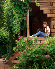 A girl sitting on a wooden building or porch and reading a book in a garden.
