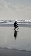 Lone cyclist riding along a reflective beach at low tide in Portugal, with ocean waves and a peaceful, moody atmosphere