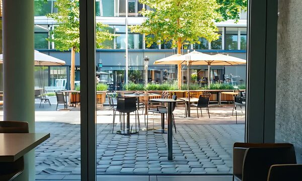 Sunlit Courtyard View through Glass Doors Featuring Tables Chairs and Umbrellas Framed by Greenery and Buildings With