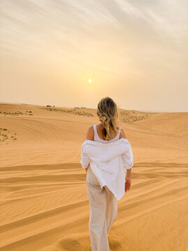 Woman walking on sand dunes in the desert towards the setting sun, facing backwards to the camera.
