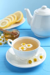 Delicious chamomile tea in cup, fresh flowers, lemon and honey on light blue wooden table, closeup