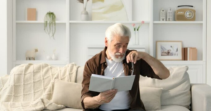 Senior man with vision problem reading book on sofa at home, camera moving down