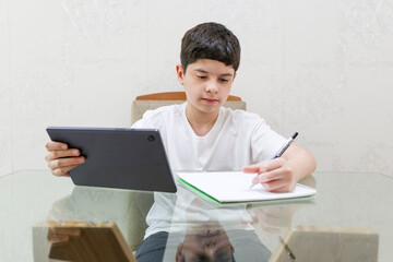 Brazilian preteen boy holds a tablet while writing in his notebook. He is left-handed, wears a white shirt, and is seated at a table indoors. His eyes are focused on the notebook