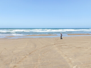 On a breezy spring day in May by the Sea of Japan, a Japanese man in his late seventies walks alone on the beach, captured from above in an aerial photo showing waves and quiet solitude.