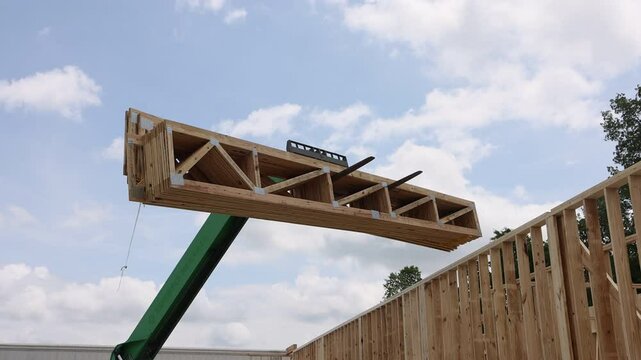 Construction site forklift telehandler lifting wooden trusses beams into place under clear blue sky.