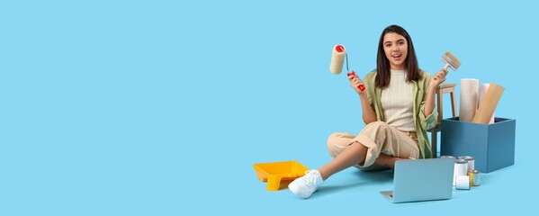 Young female interior designer with paint roller, brush, laptop and wallpapers on blue background...