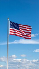 Fototapeta premium An American flag waving gracefully against a bright blue sky with scattered, gentle white clouds.