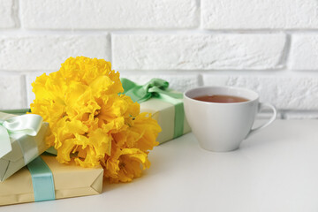 Narcissus flowers with gifts and cup of tea on table near white brick wall