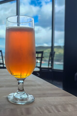 A pint glass filled with a pale ale craft beer. The liquid has a yellow tint. A clear beer glass sits on a table at a microbrewery. The background is the bar with taps and glasses. 