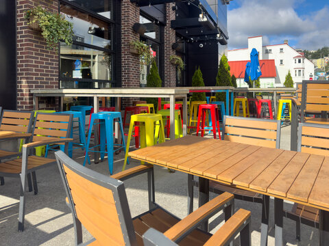 Colorful iron metal stackable bar stools, blue, red, and yellow in color. The cafe chairs are next to rows of long wooden pub tables on the outside of a restaurant in the patio garden.