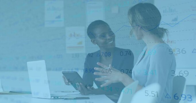 Two women sharing tablet while starting finance review, one gesturing to graph and typing on laptop