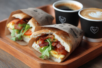 Delicious egg and bacon sandwich wrapped in paper, served with coffee on wooden tray