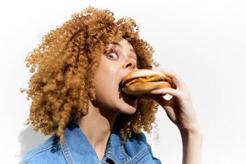 Young woman with curly hair excitedly eating a cheeseburger against a plain white background, showcasing a vibrant and playful food moment