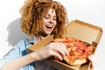 Young woman with curly hair joyfully holding a pizza box, wearing casual attire, against plain background, embodying happiness and a love for comfort food