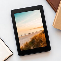 A black e-reader displaying a serene beach landscape with dunes and a calm sea at sunset is positioned on a white surface next to a stack of books.