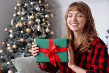 Happy young woman in pajamas with Christmas gift sitting at home