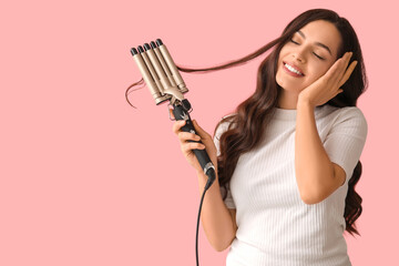 Beautiful young woman with curling iron on pink background